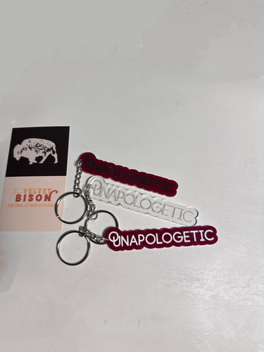 A red and clear plastic keychain with the word 'Unapologetic' printed on it, attached to a metal keyring.